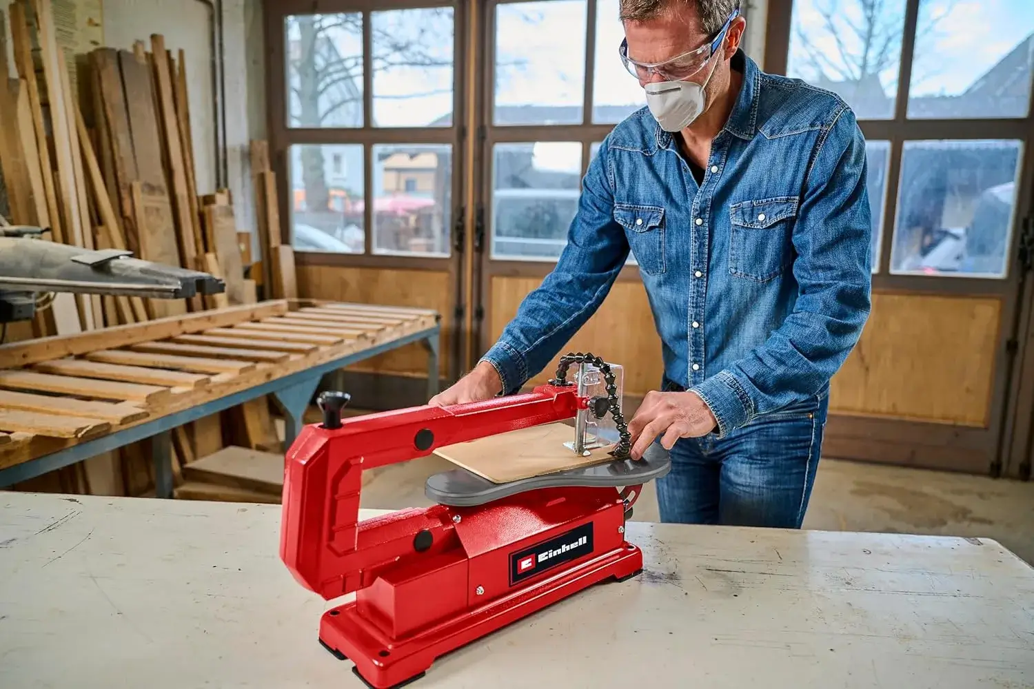 Ein Handwerker in einer Werkstatt nutzt eine rote Einhell Dekupiersäge, um präzise Holzarbeiten durchzuführen. Im Hintergrund sind Holzmaterialien und Werkstattelemente zu sehen. Ideal für DIY-Projekte und feine Sägearbeiten.